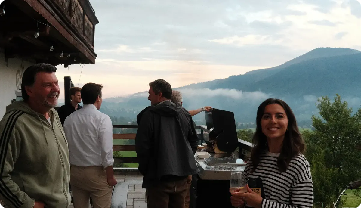 Group of people socializing on a balcony with a scenic mountain view. A woman in the foreground smiles at the camera.