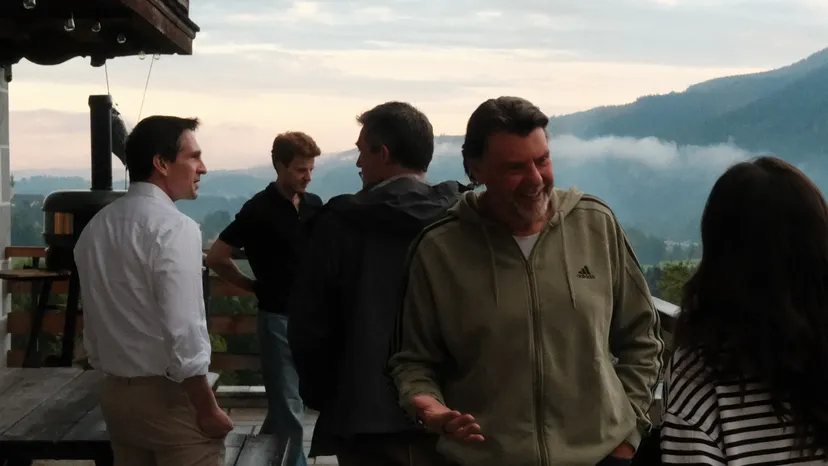 A group of people chatting on a balcony with a scenic mountain view in the background. One person is wearing a green hoodie.
