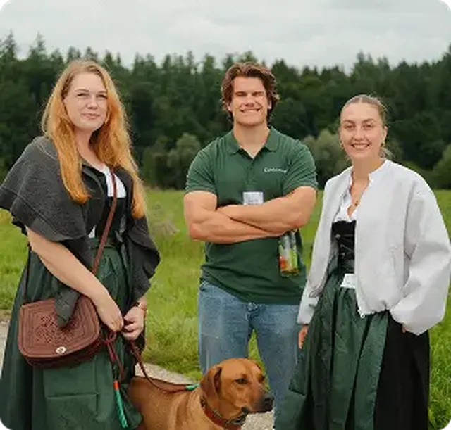 Three people and a dog standing outdoors. Two women in dresses and a man in a green shirt smile, with trees in the background.