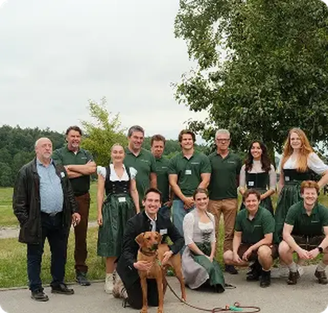 A group of people, some in traditional attire, pose outdoors with a dog. Trees and a cloudy sky are in the background.