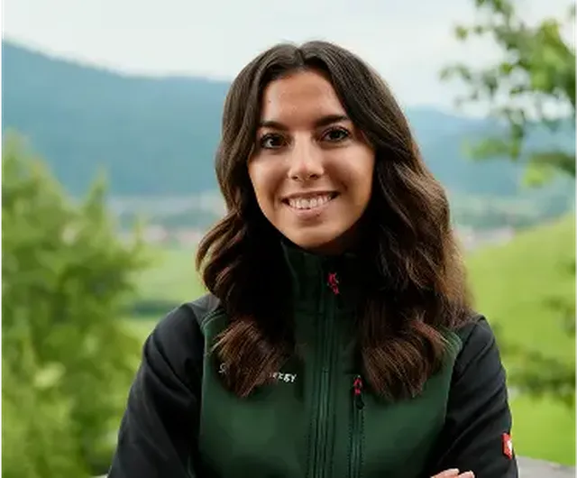 A person with long dark hair smiles, standing outdoors in a green jacket. The background shows blurred greenery and hills.