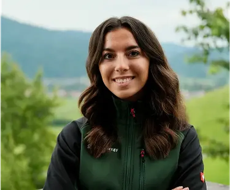 A person with long dark hair smiles, standing outdoors in a green jacket. The background shows blurred greenery and hills.