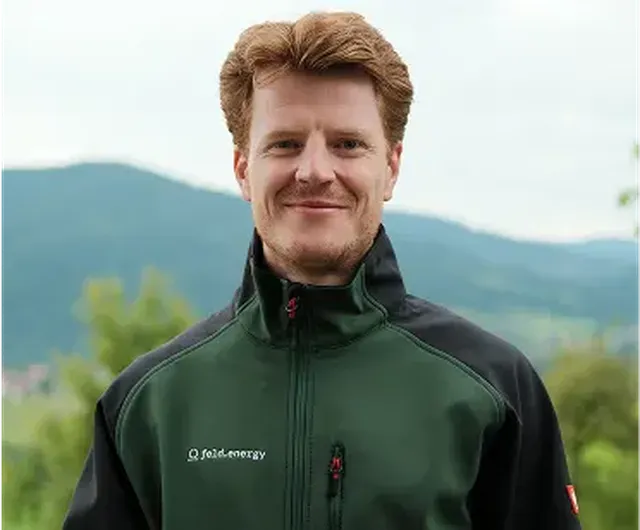 A person with red hair in a green and black jacket stands outdoors with hills and trees in the background.