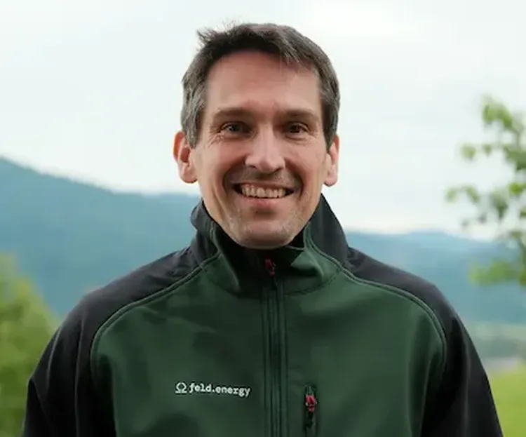 Man smiling outdoors in a green jacket with "field.energy" logo, mountains and trees in the background.