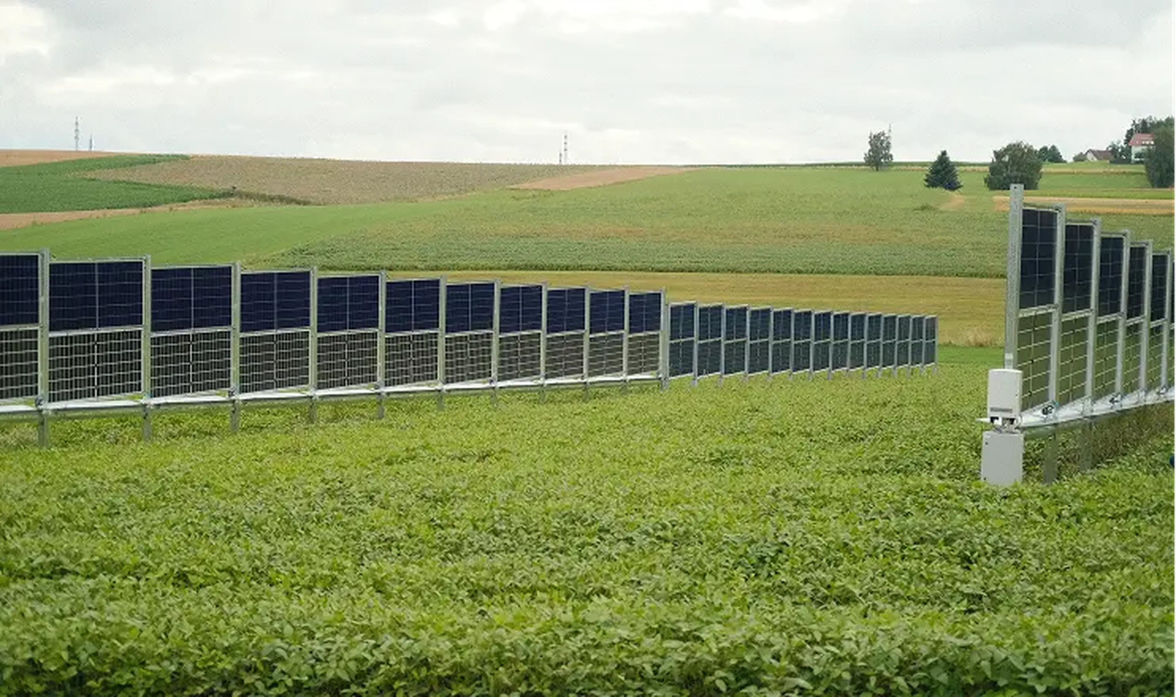 Rows of solar panels in a lush green field under a cloudy sky, with rolling hills in the background.
