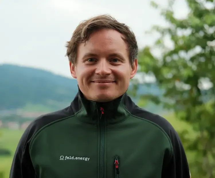 A person wearing a dark green jacket stands outdoors, smiling, with blurred greenery and hills in the background.