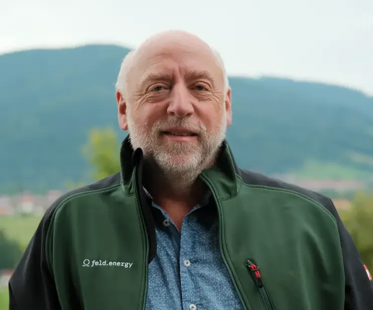A man with a beard wearing a green jacket stands outdoors with hills in the background.