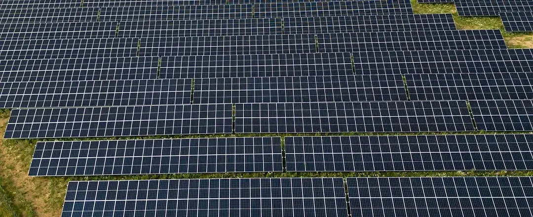 Aerial view of a large solar panel farm with rows of dark panels on a grassy field.
