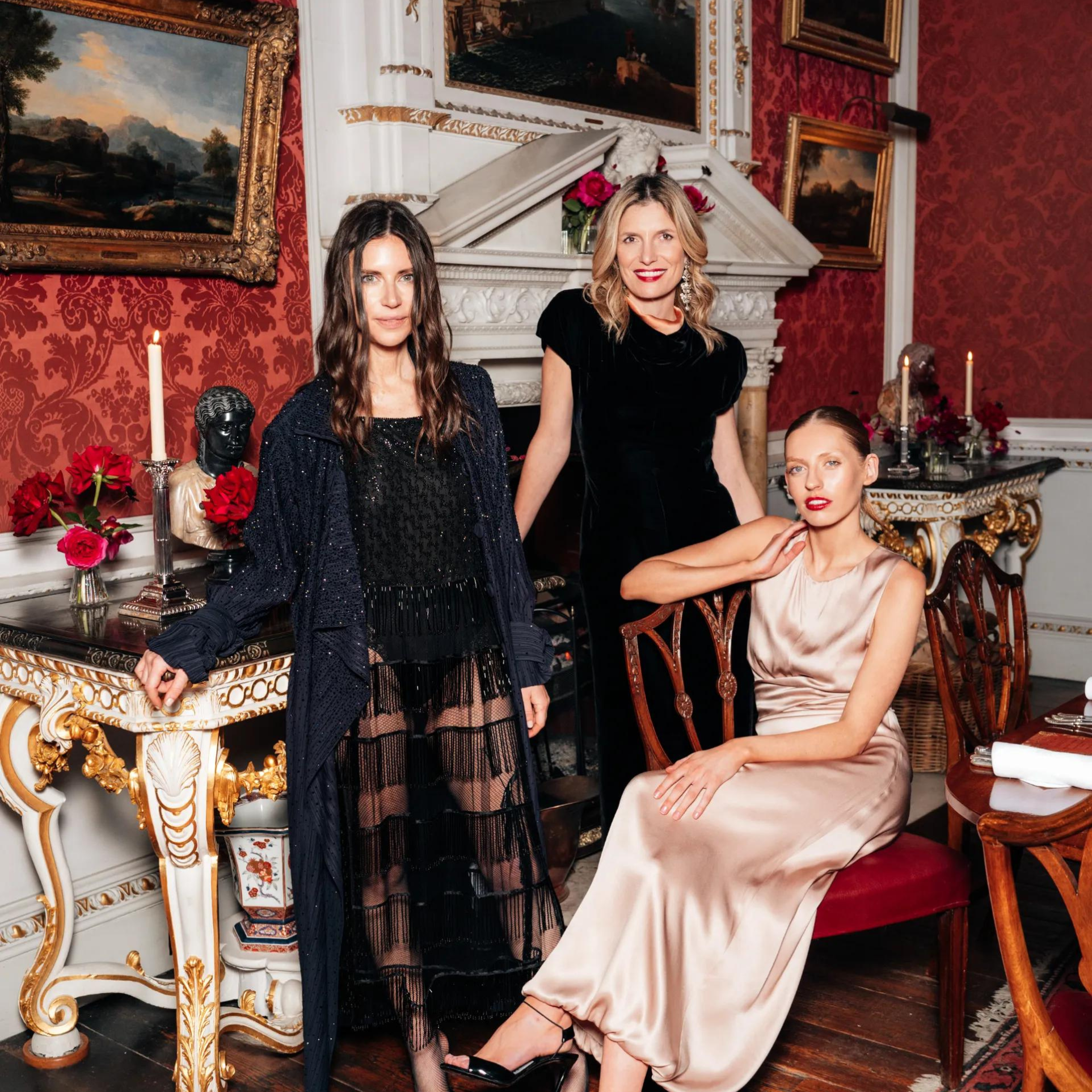 Three elegantly dressed women with polished hair and makeup at an event, in a richly decorated room with red damask walls, gilt-framed paintings, and candlelight.