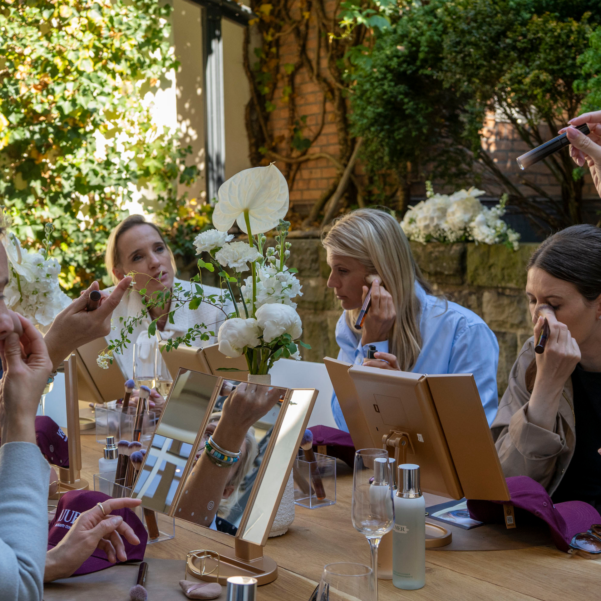 Guests receiving makeup applications at a Ruuby beauty event, seated at a flower-dressed table with lit vanity mirrors in a garden setting