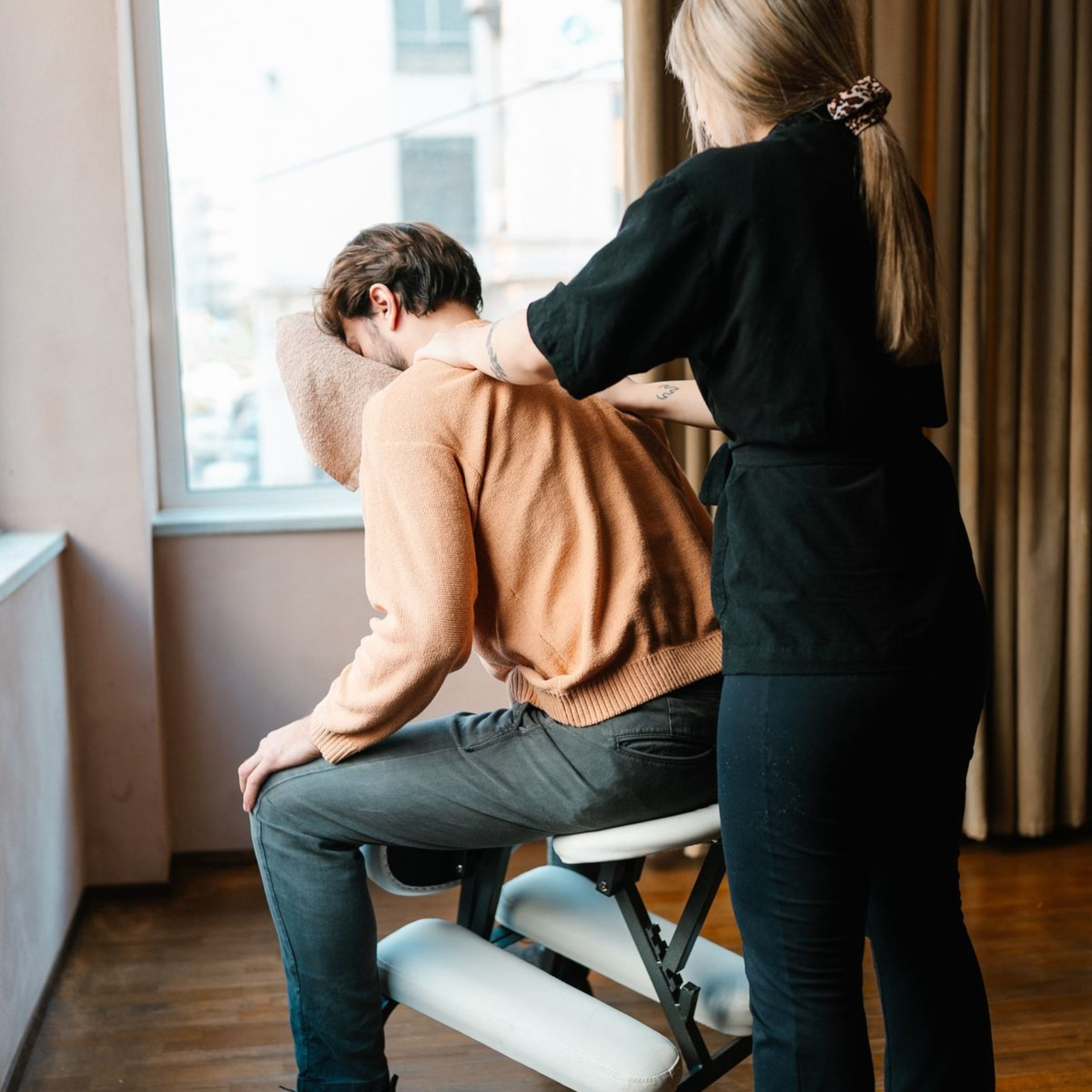 A therapist giving a man a seated chair massage, working into his shoulders as he rests forward on the cushioned headrest by a window.