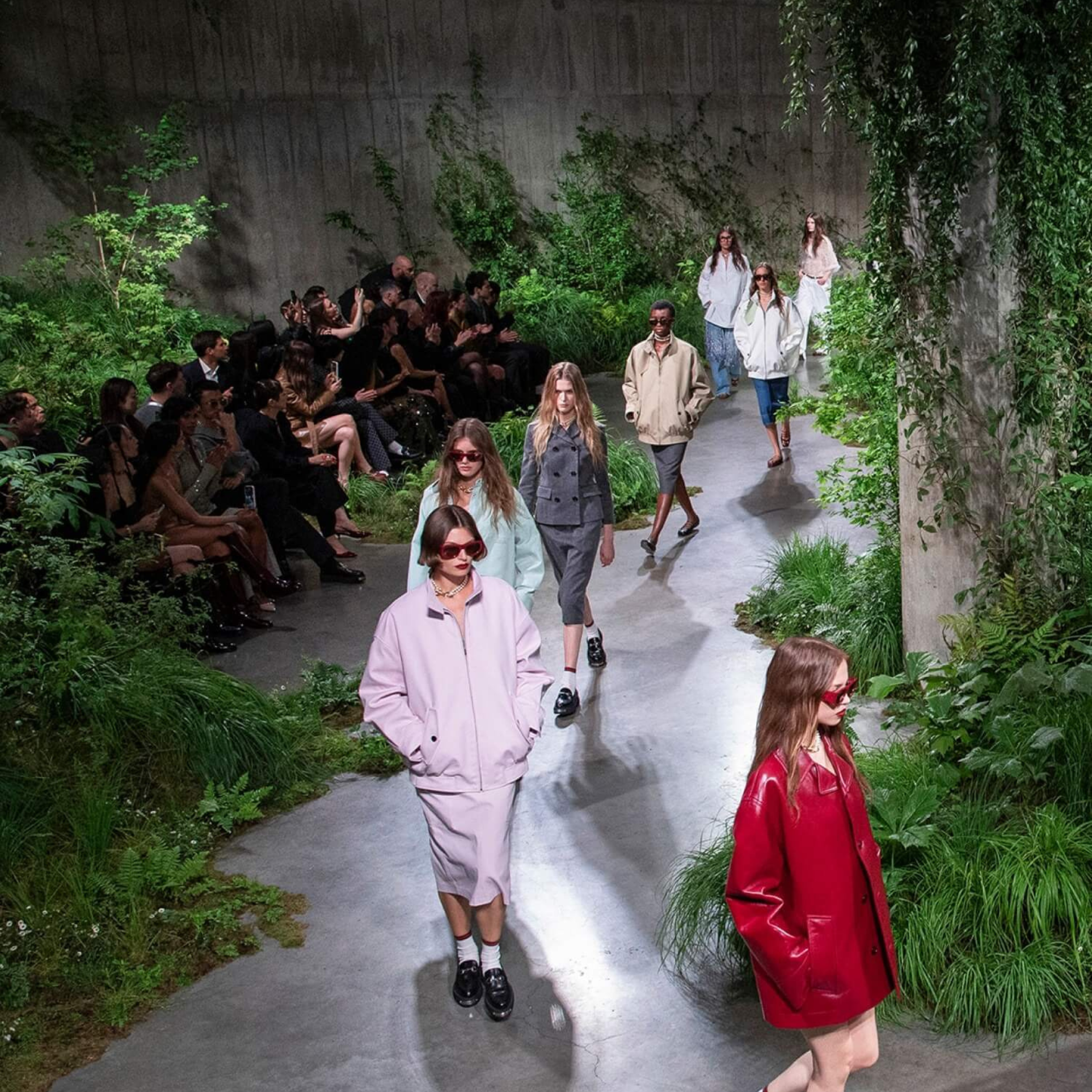 Models walking the runway through a greenery-lined set at a fashion show, with guests seated front row