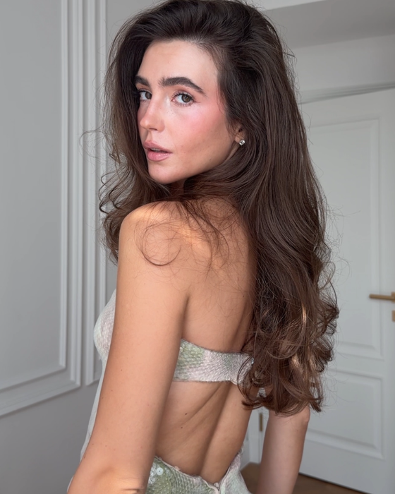 Tetiana Soviak looking over her shoulder with long, voluminous wavy brown hair and natural glowing makeup, wearing a strapless top in a softly lit room