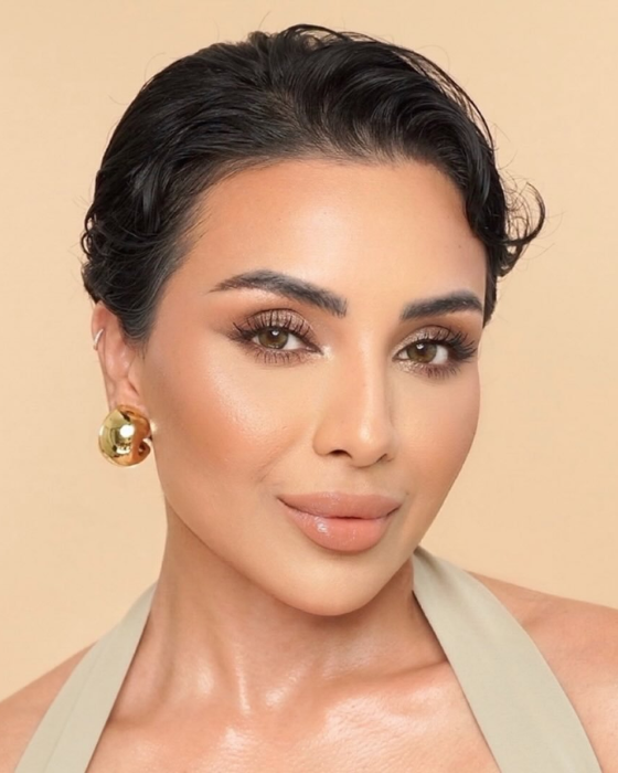 Nina Ubhi in a close-up beauty portrait with slicked-back dark hair, soft glam makeup and a gold hoop earring, against a beige background.