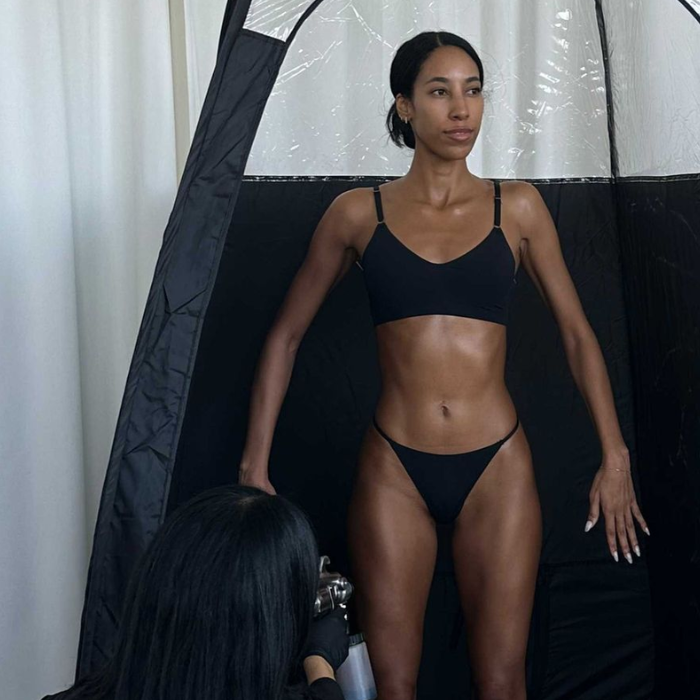 Woman standing in a pop-up tanning tent with a contoured, sculpted spray tan, as a tanning expert works on her lower body.