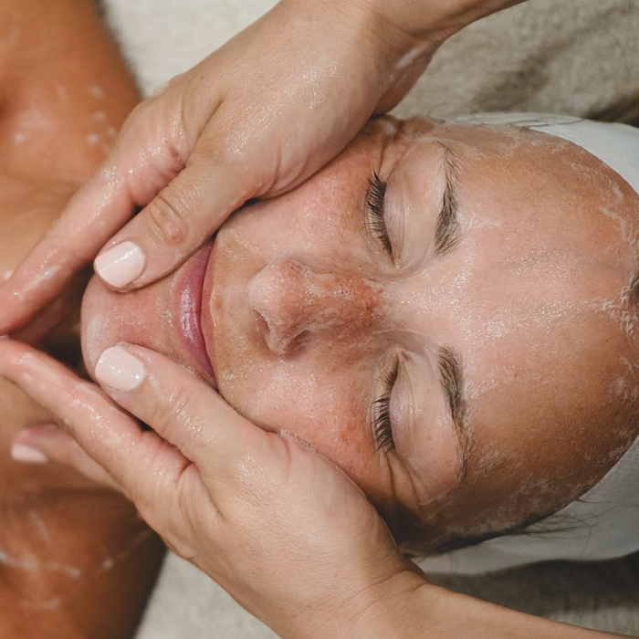 Facialist performing a bespoke facial, massaging cleanser or serum into a woman's face as she relaxes with her eyes closed.