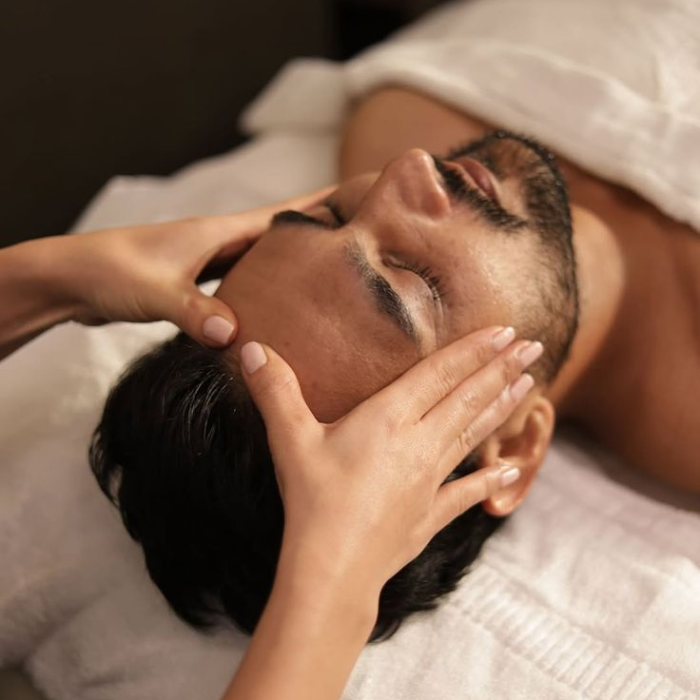 Facialist performing a facial massage on a man's face as he relaxes with his eyes closed on a treatment bed.