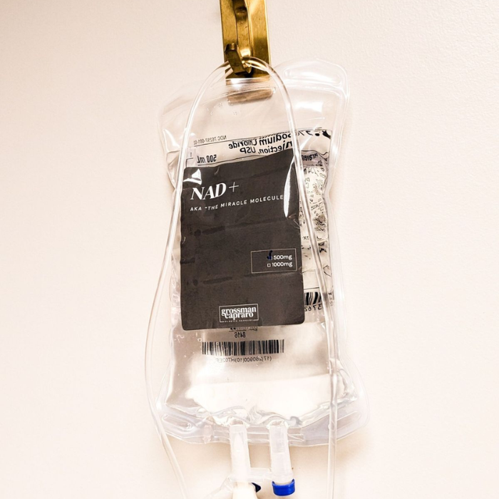 A clear NAD+ IV drip bag with a black label, hanging from a gold clip against a white wall.