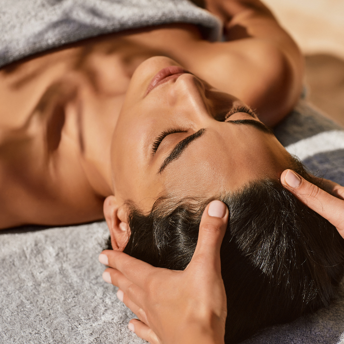 A therapist's hands performing a L'Occitane en Provence Aromachology massage on a client's head and scalp, as she relaxes with her eyes closed.