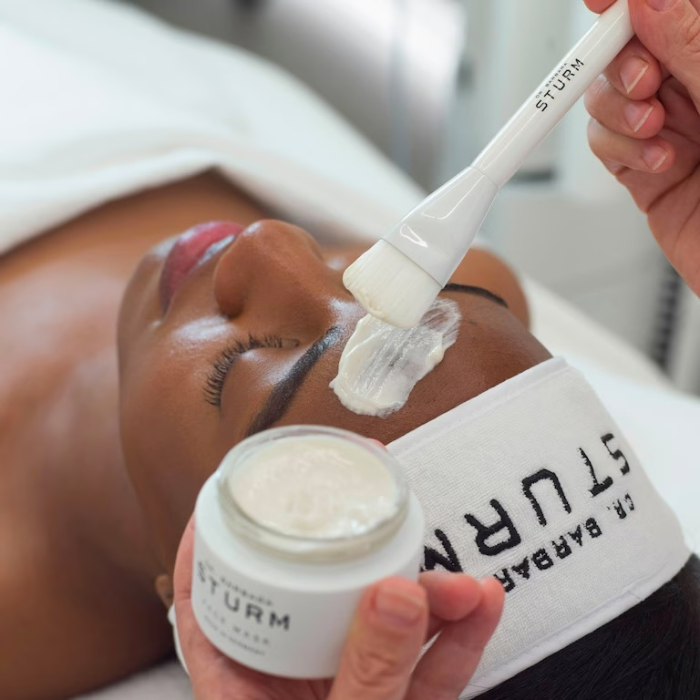 Claude responded: Facialist applying a Dr.Facialist applying a Dr. Barbara Sturm face mask to a woman's forehead with a branded brush, with the product jar and headband in view.