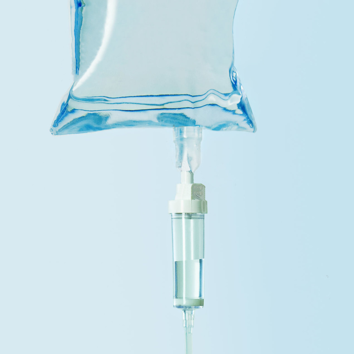 A clear IV drip bag and attached flow chamber against a pale blue background.