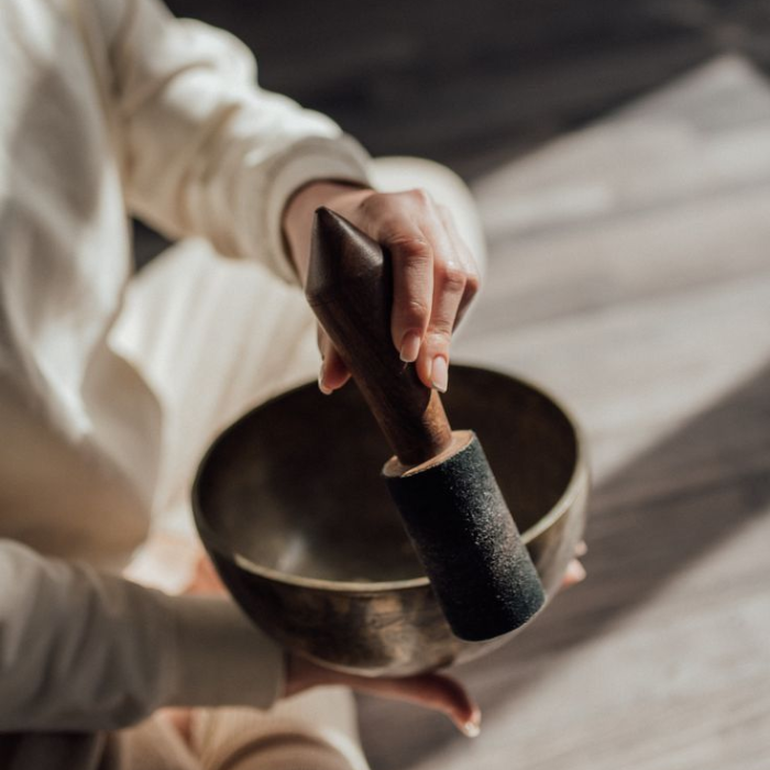 A person in white clothing playing a Tibetan singing bowl with a wooden mallet, seated on a wooden floor in soft light.