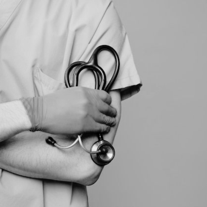 A medical professional in scrubs with arms crossed, holding a stethoscope, shown from the shoulders down against a grey background.