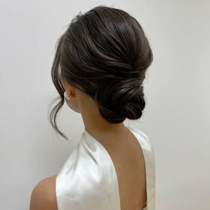 Back view of a person with dark, glossy hair styled in an elegant low twisted bun. Soft face-framing curls fall near the cheeks, and the hairstyle is paired with a white satin halter-style garment featuring an open back. The image is set against a plain light-coloured background, highlighting the sleek, polished updo.