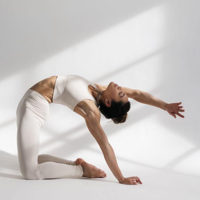 A woman in cream activewear performing a kneeling yoga backbend in a bright, sunlit white room.