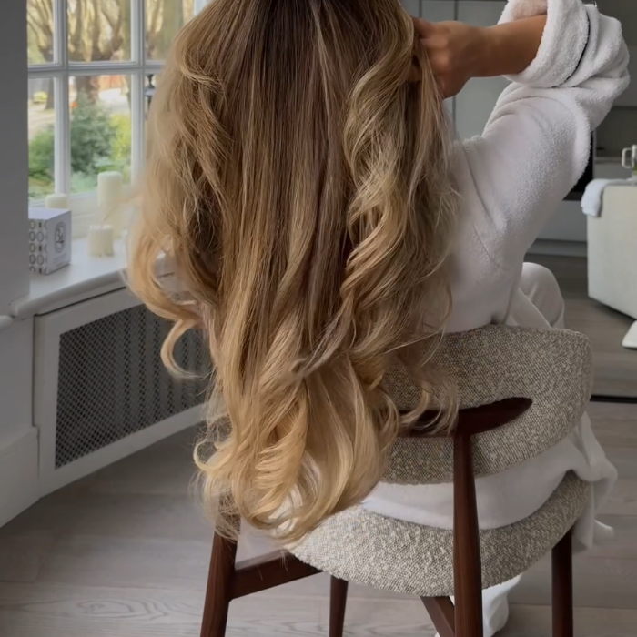 Long blonde hair styled in soft waves after an at-home blow-dry, shown from behind by a sunlit window
