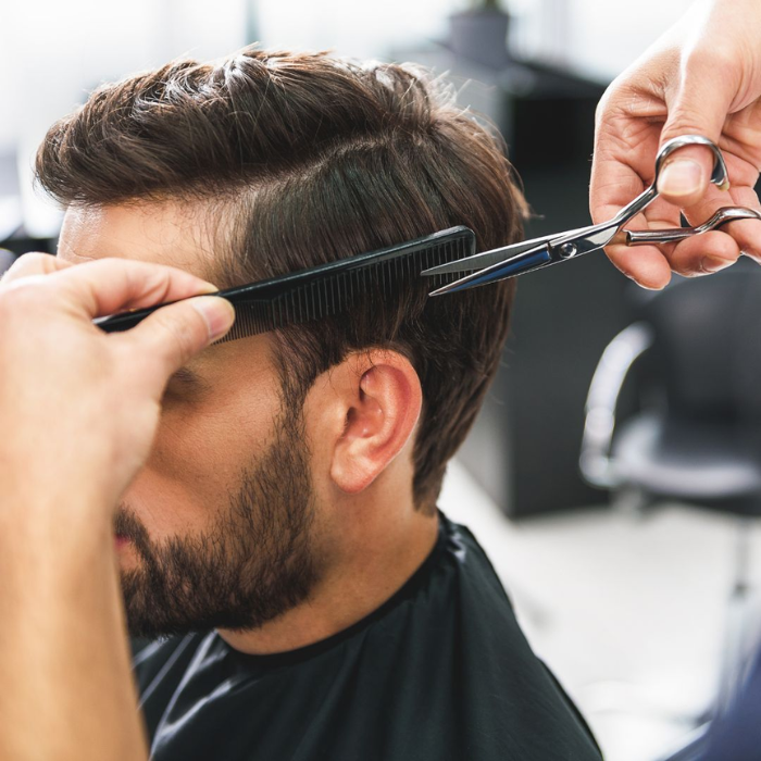 Hairdresser cutting a bearded man's hair with comb and scissors, working through the top section.