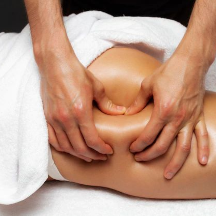 A therapist's hands performing a firm anti-cellulite massage on a person's thigh, draped with white towels.