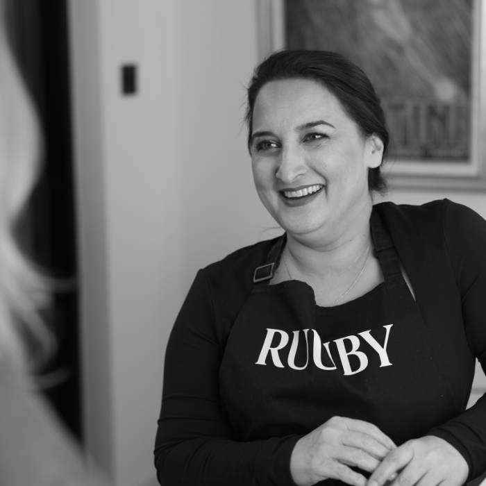 A smiling Ruuby therapist wearing a branded black apron, talking with a client, in a black-and-white photo.