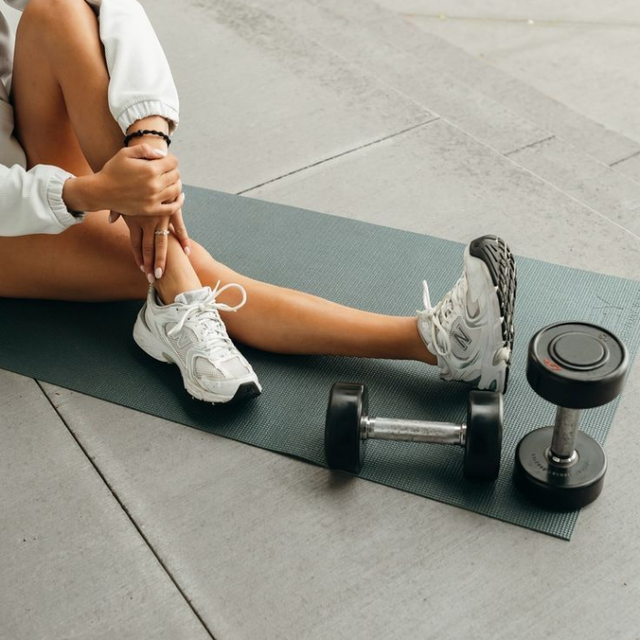 A woman in activewear and white trainers seated on a yoga mat beside a pair of dumbbells.