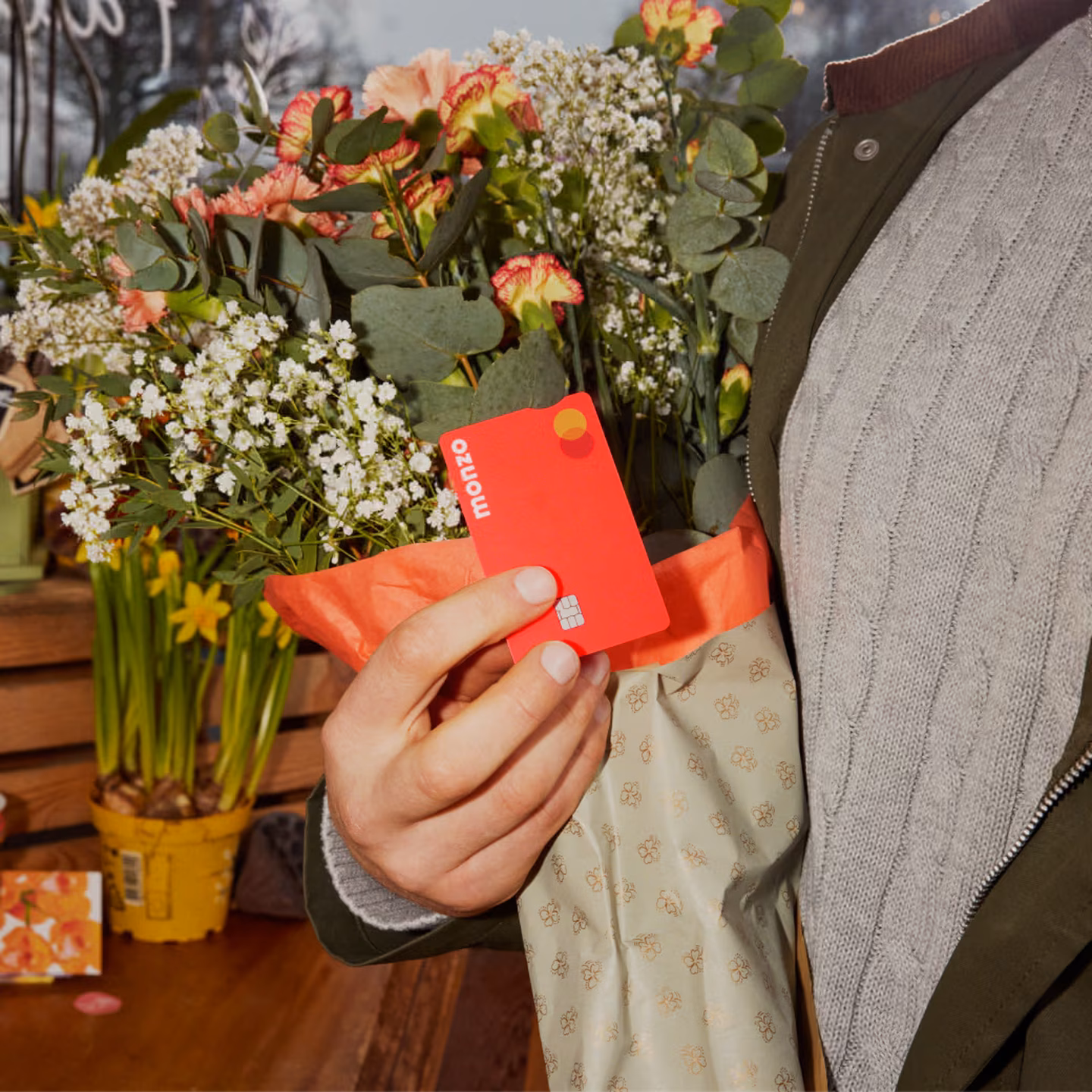 Una persona sujeta una tarjeta de Monzo de color coral enfrente de un ramo de flores frescas.