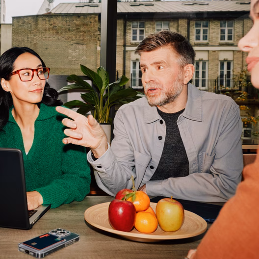 Three colleagues talking around a table with laptops during a meeting.