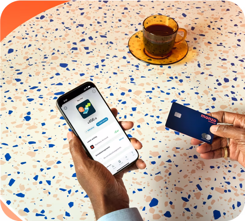 Person at a terrazzo table holding a phone showing their Monzo Business Tax Pot balance, with a Monzo business card in their other hand and a cup of tea nearby.