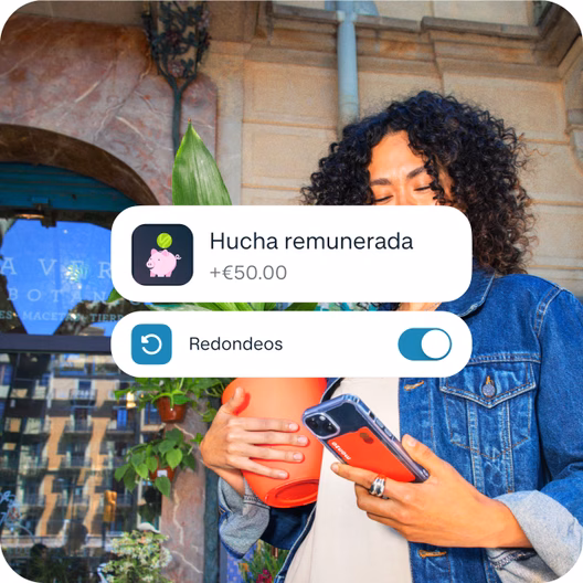 Una mujer con un móvil y una planta en maceta, con una interfaz de la app de Monzo superpuesta que muestra 50 € añadidos a una Hucha remunerada y la función de redondeos activada.