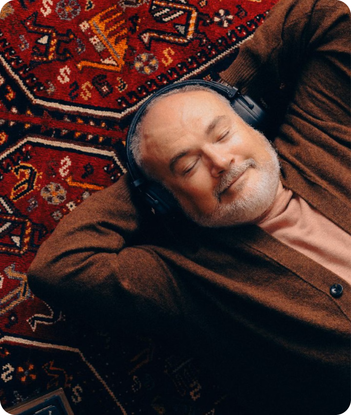 Photo of a man lying down on a red carpet, contentfully listening to music with overhead earphones. 