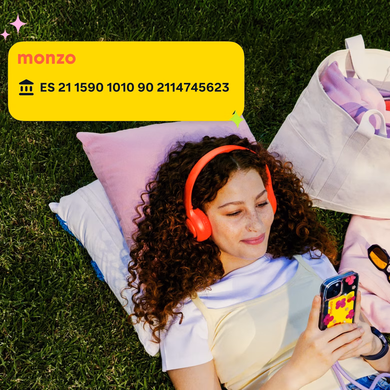 A woman lying on the grass wearing red headphones and looking at her phone, with a Monzo overlay showing a Spanish IBAN on a yellow card.