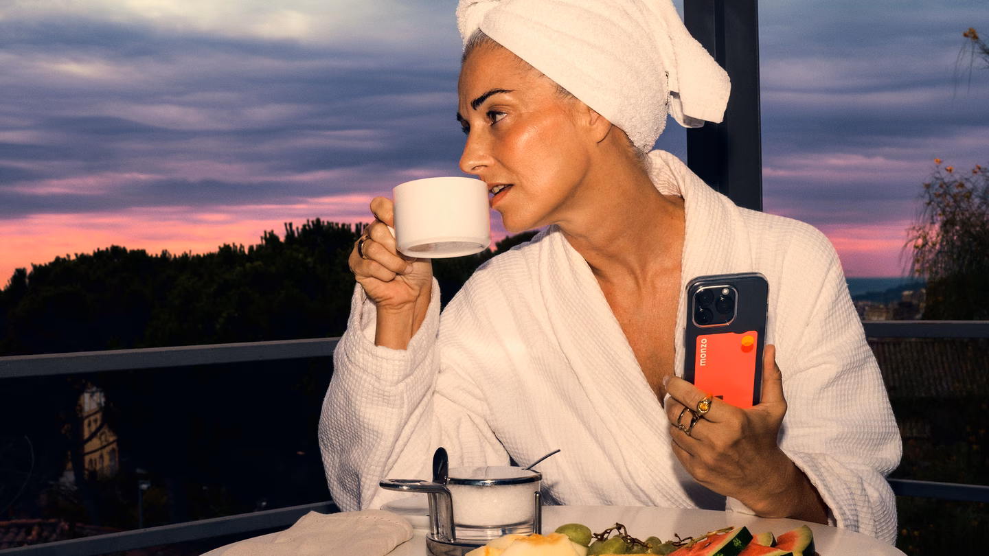 A woman in a bathrobe enjoys coffee and breakfast on a balcony at sunset, holding a phone with a Monzo card in the case