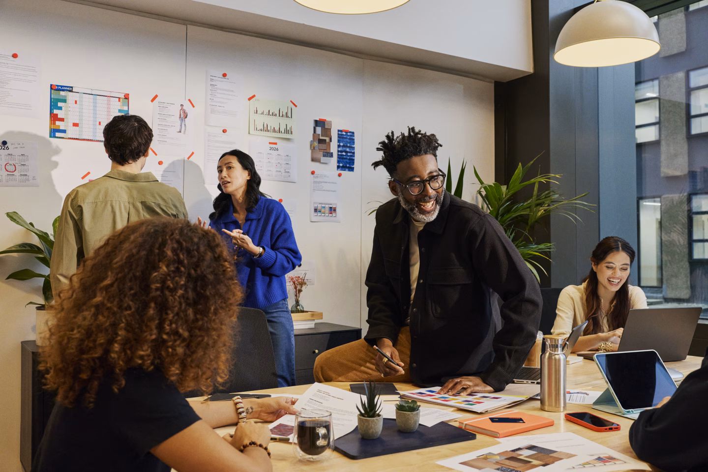 A diverse team collaborates around a table in a bright modern office, reviewing documents and charts. The workspace features pendant lighting, large windows, and a wall covered with planning materials and data visualizations.