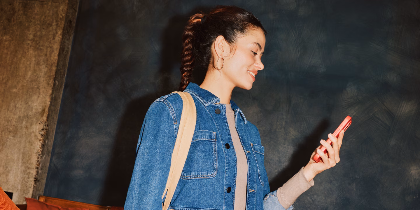 Woman in a denim jacket smiling at her phone in a coral case.