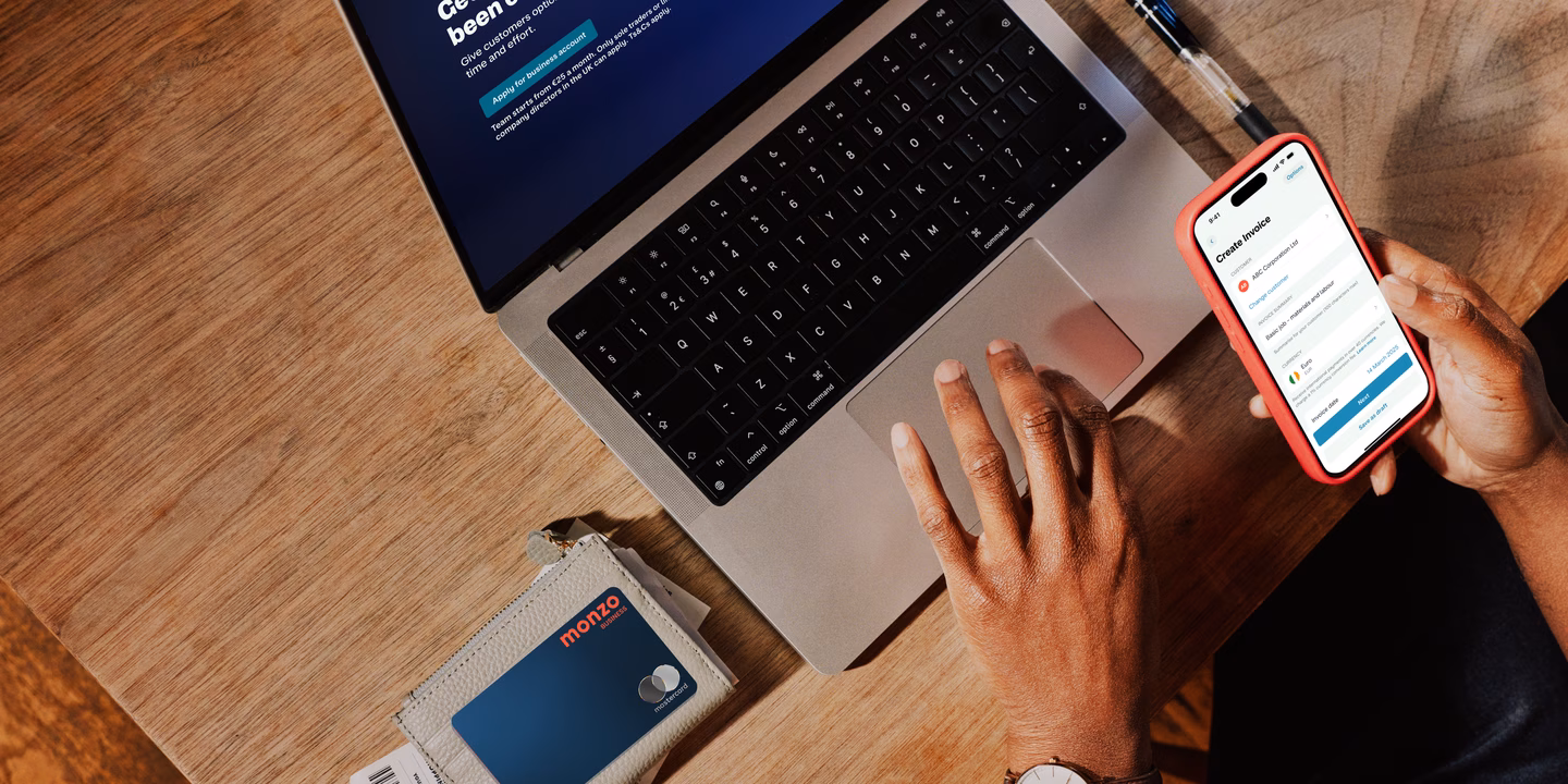 Overhead view of a wooden desk with an open laptop showing the Monzo Business dashboard, one hand on the trackpad and the other holding a phone in a coral case displaying a "Create invoice" screen, with a Monzo card in a cardholder nearby.