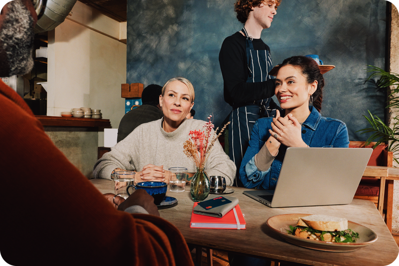 People meeting around a café table with a laptop and coffees, with an aproned café worker nearby.