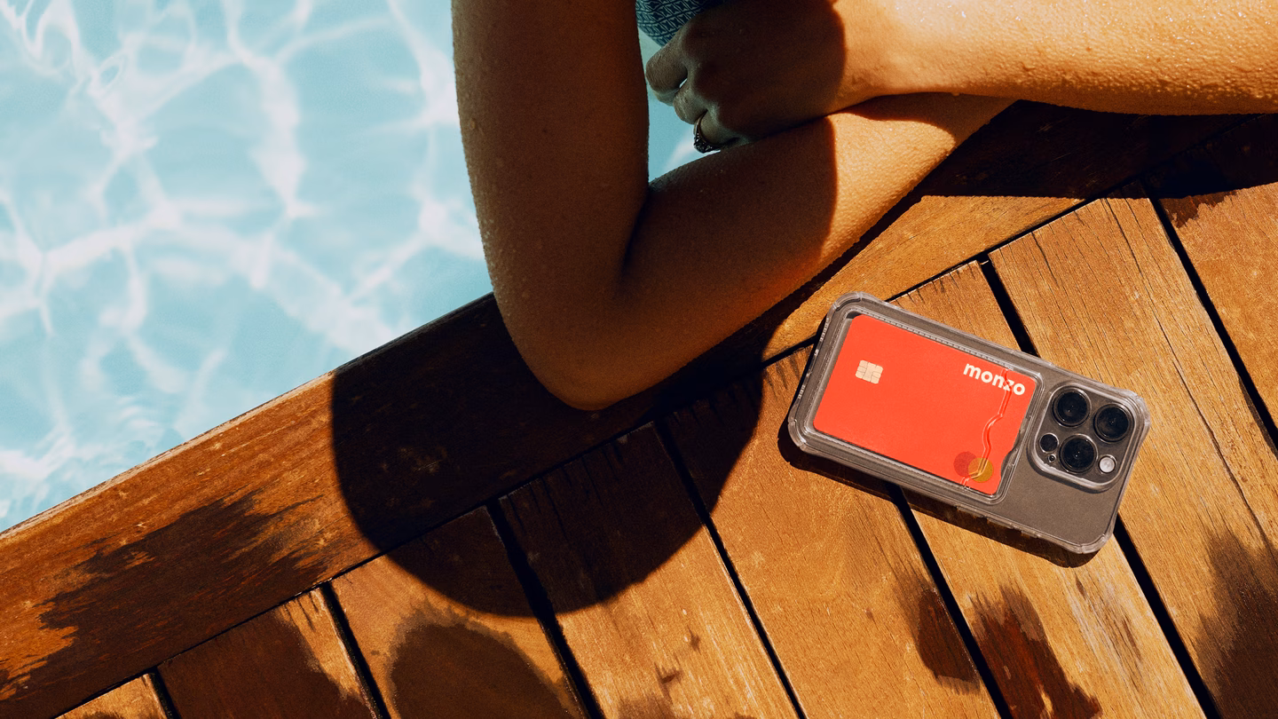 A person relaxing by a sunlit swimming pool, with a coral Monzo card tucked into a phone case on the wooden deck beside them.