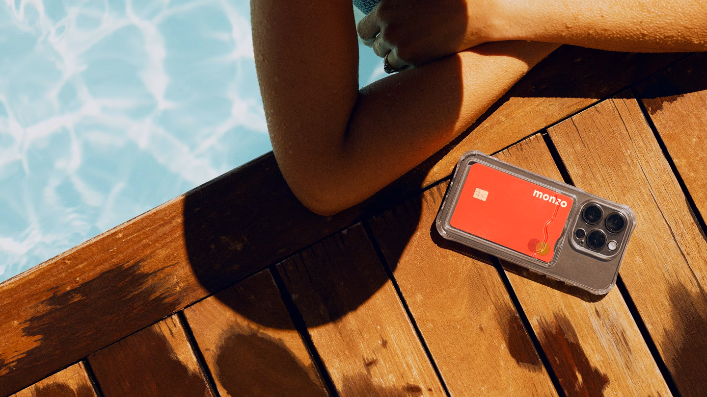 Una persona descansa en una piscina bañada por el sol y, a su lado, encima del suelo de madera, hay una tarjeta de Monzo de color coral dentro de una funda de móvil.