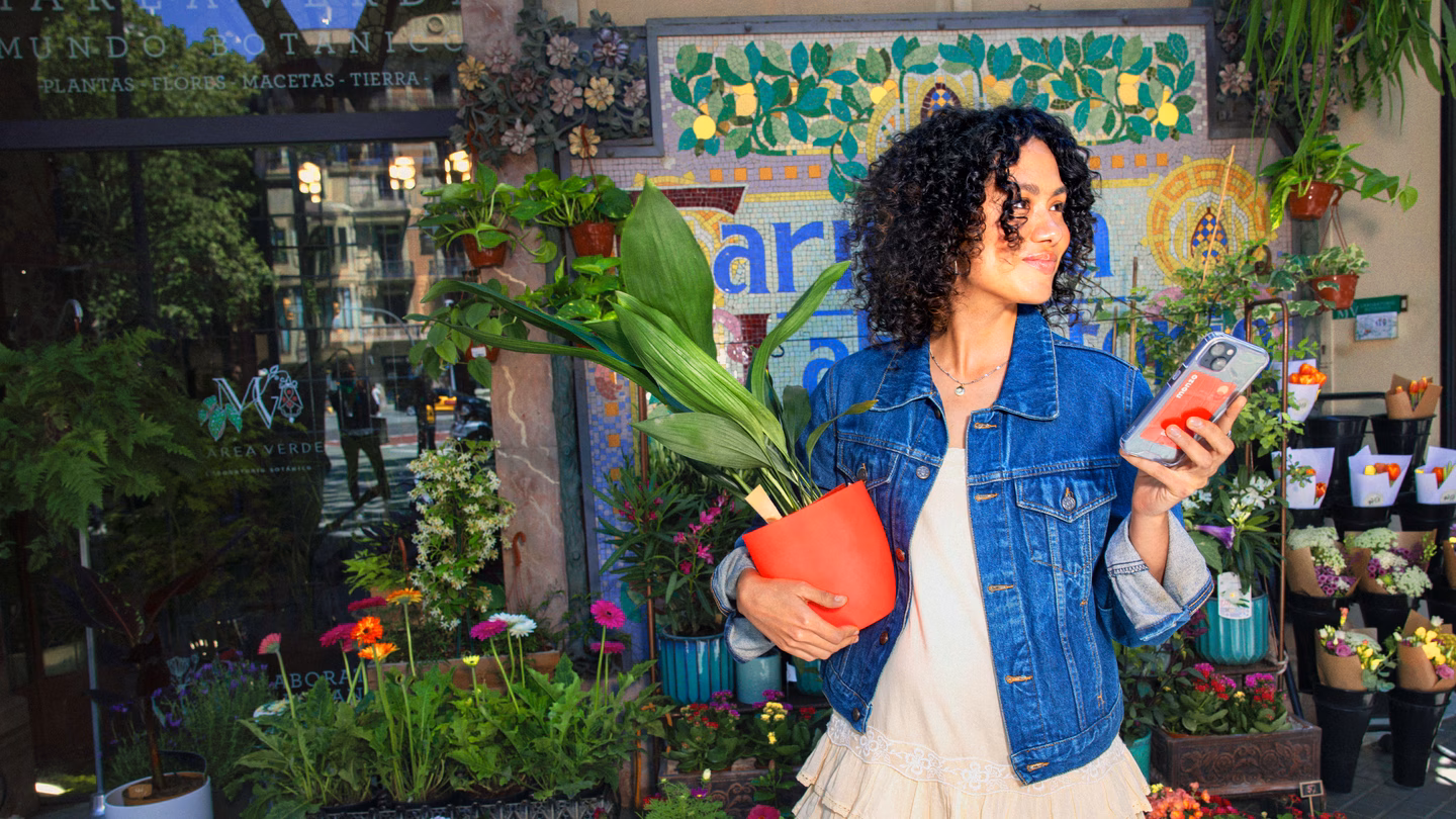 Mujer con flores, una barra de pan y una tarjeta de color coral de Monzo paseando por un colorido mercado español.