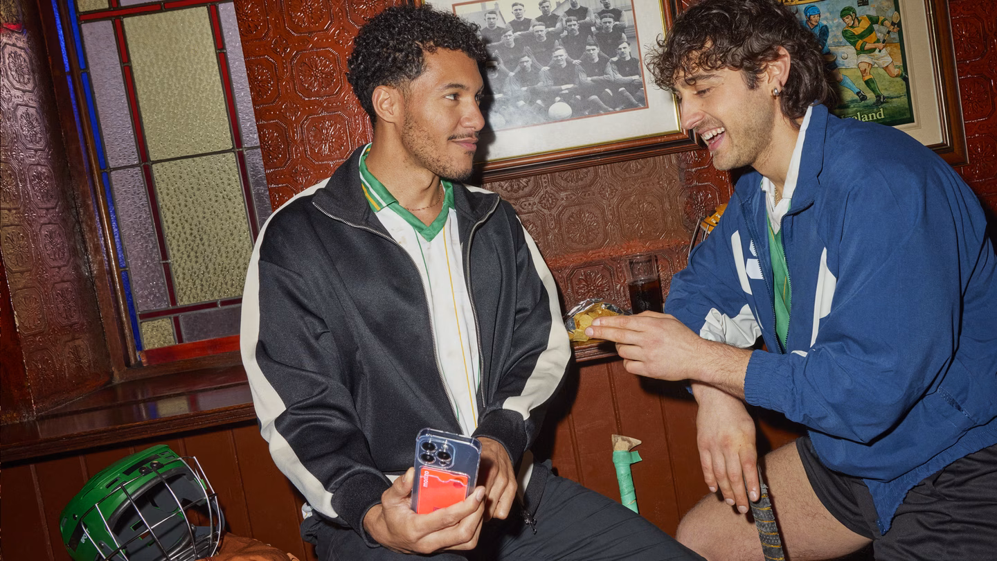 Two friends chatting in a traditional pub with vintage football memorabilia on the walls. One holds a phone while the other holds a drink, sharing a lighthearted moment.