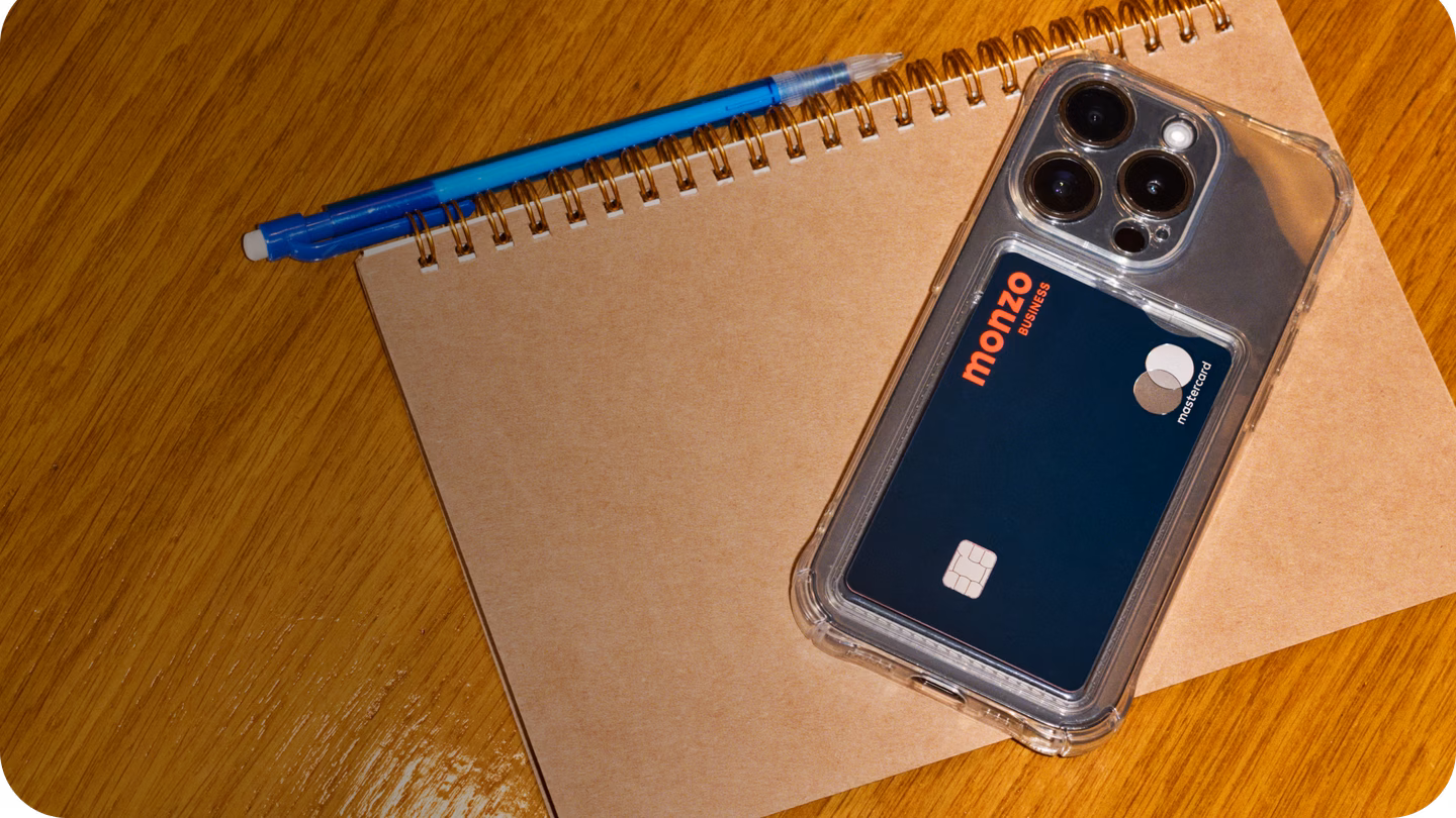 A navy Monzo Business card in a phone case resting on a notebook and pen on a wooden desk.